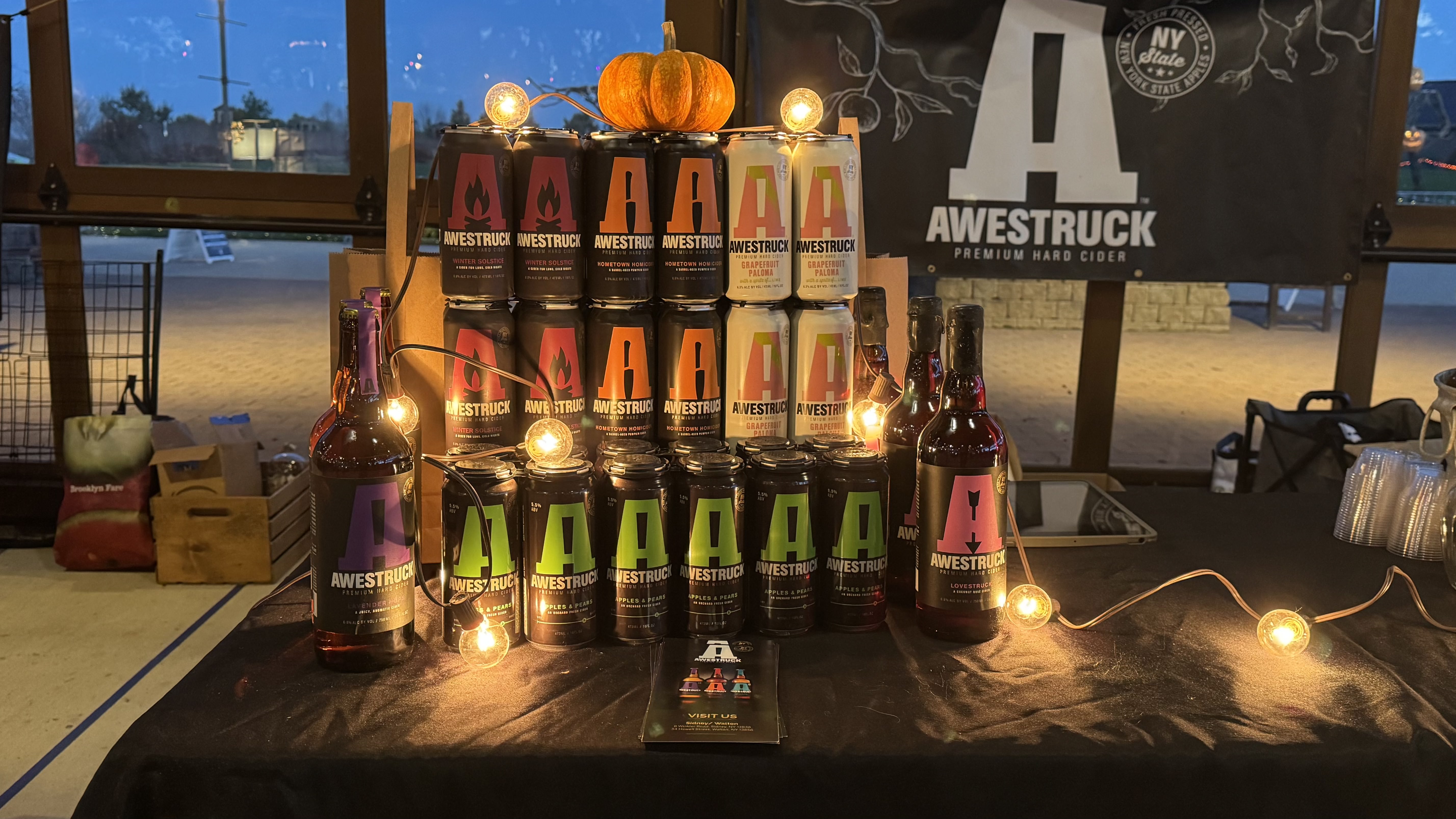 Display of 4 canned ciders (Hibiscus Ginger, Peach Ring, Honeycomb, and Summer Sangria) outdoors against a sunset background